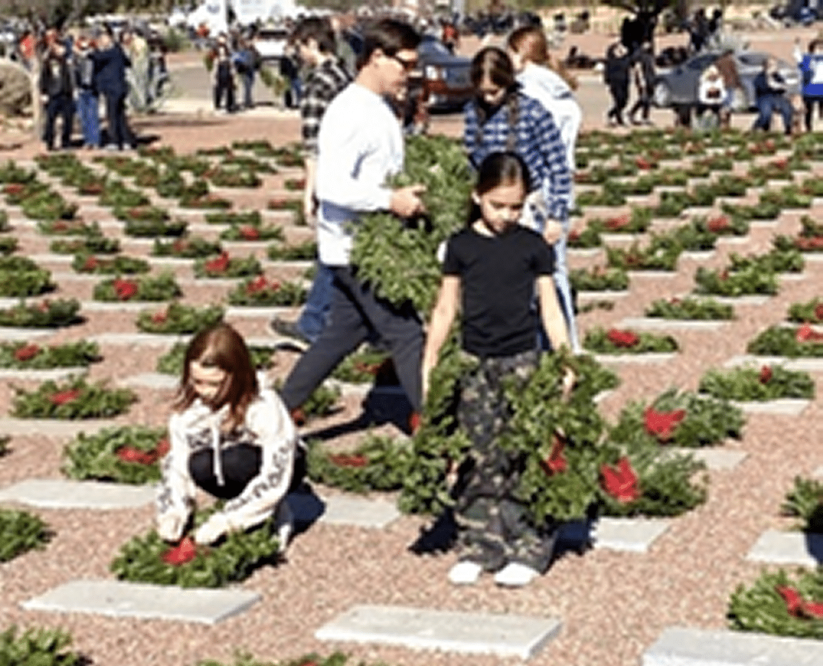 Young ladies and men placing green wreaths with red ribbons on headstones at Veterans cemetery with hundreds of wreaths and headstones in the background.