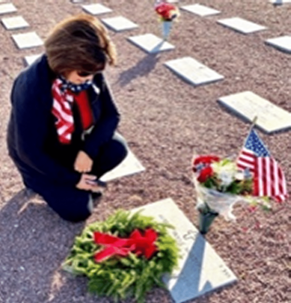 Lady in dark blue kneeling by gravestone after placing a green wreath with a red ribbon on the gravestone.