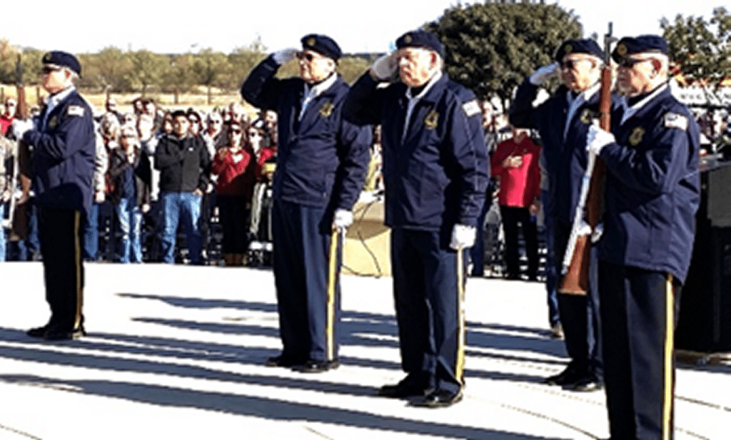 Four members of Post 132 Honor Guard in dark blue uniforms saluting with a crowd of attendees in the background.