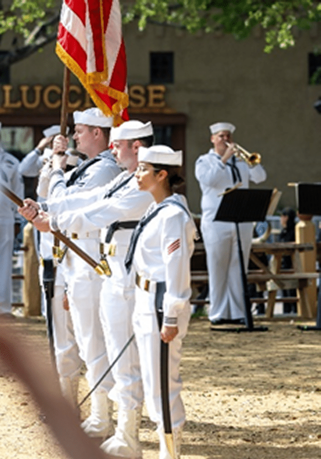 Four U. S. Navy Sailors in white uniforms holding United States flag with another Sailor in the background blowing a bugle.