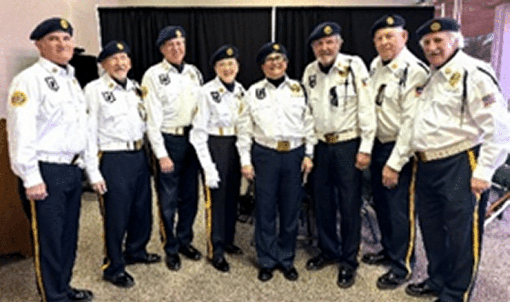 Eight members of American Legion Post 132 Honor Guard lined up in blue berets, white shirts and blue pants with yellow side stripes.