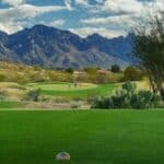 View of Golf course fairway and putting green with three golfers on green, prickly pear cactus in foreground, mountains in background under a blue sky with white puffy clouds.