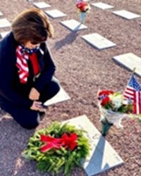 Kneeling lady placing Wreath on headstone with other headstones in background.